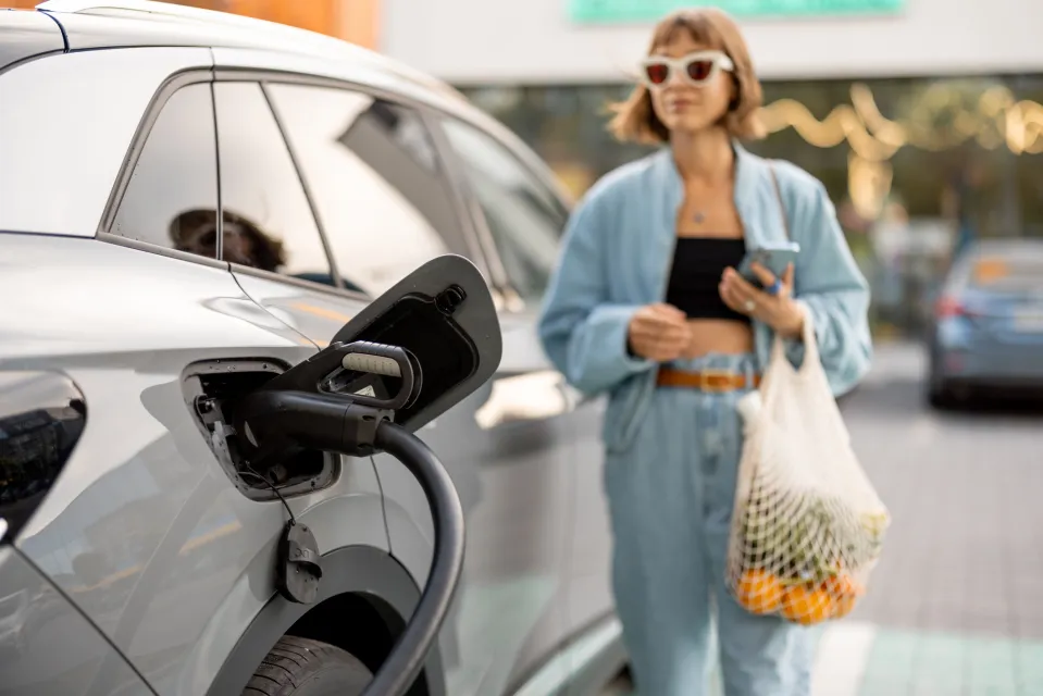 Close-up of fast charger plug in vehicle charging port, woman with groceries waiting for her car to be charged on background