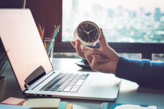 Hand holding a small clock beside an open laptop computer.