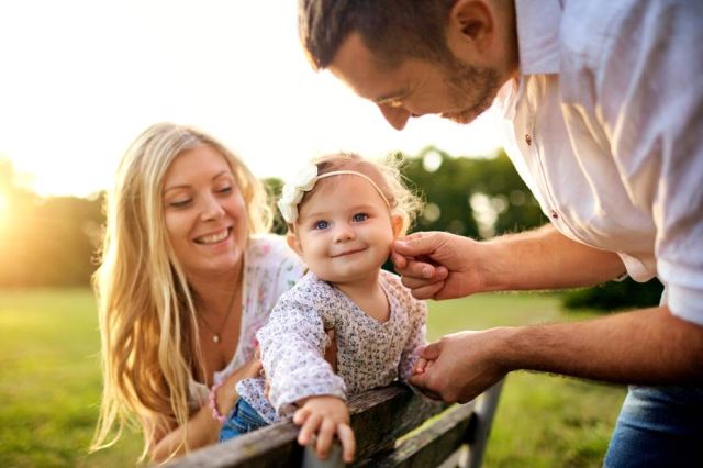 A picture showing a young happy family sitting on a bench in the park, with mom holding the baby girl and dad touching the baby's face.