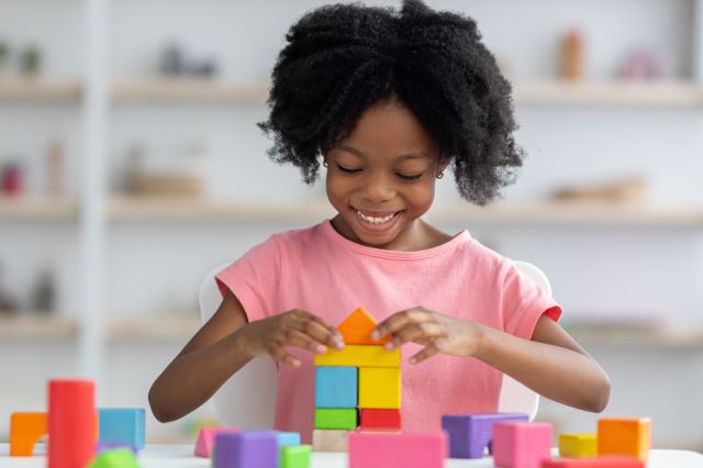 Happy young African American girl in bright pink shirt, playing with colorful wooden blocks