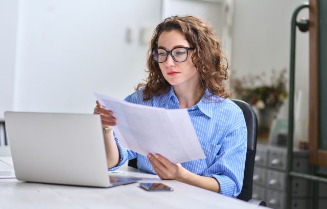 A woman looking at documents while working on a laptop