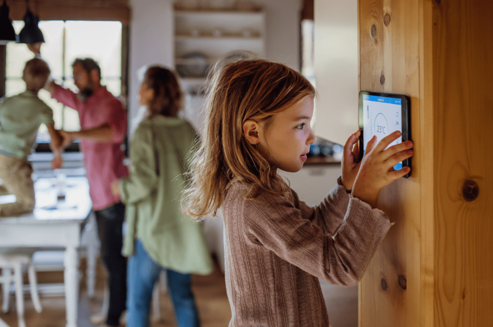 A young girl sets the temperature on a smart thermostat with her family at home