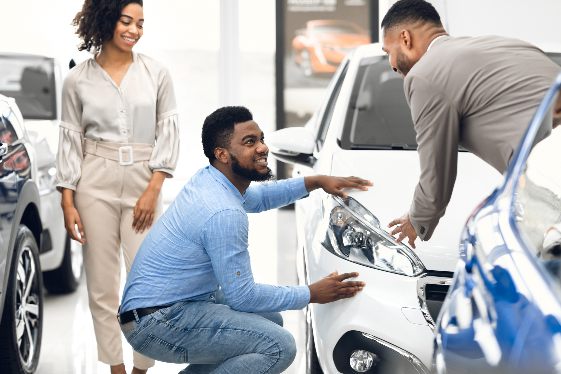 As his wife stands next to him, a man crouches beside a white car at a dealership, inspecting the front of the car while speaking to a dealer.