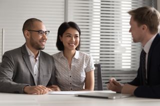 Smiling newcomer couple meeting with a banker to create a financial plan. The article highlights various ways newcomers to Canada can save for a down payment to buy a home.