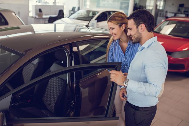 Male car dealer holds open the door of a small, grey car and explains its features as a smiling female car buyer peers in to get a look at the car's interior