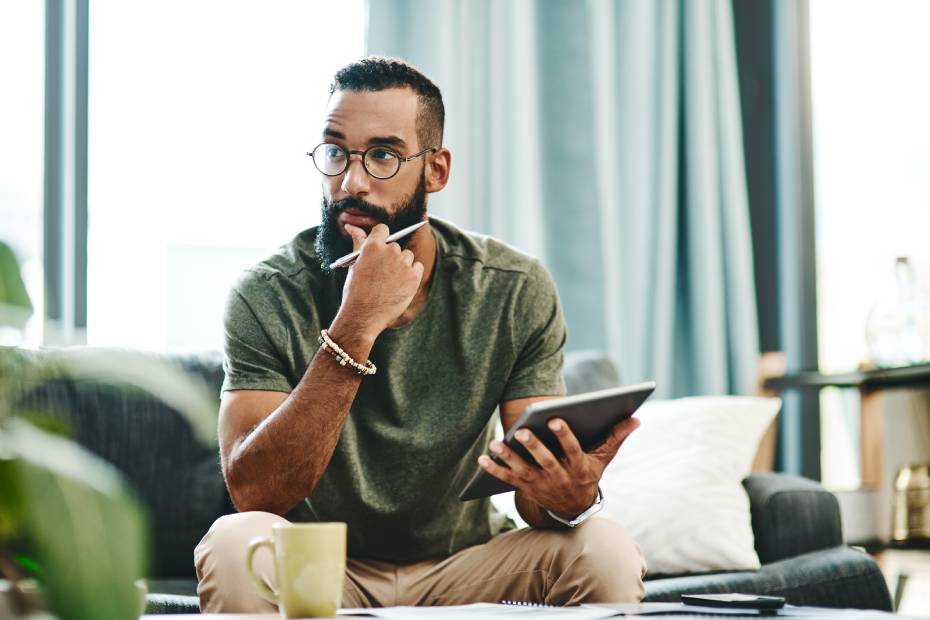 Image d'un jeune homme pensif assis sur un sofa, qui tient un crayon et une tablette dans les mains.