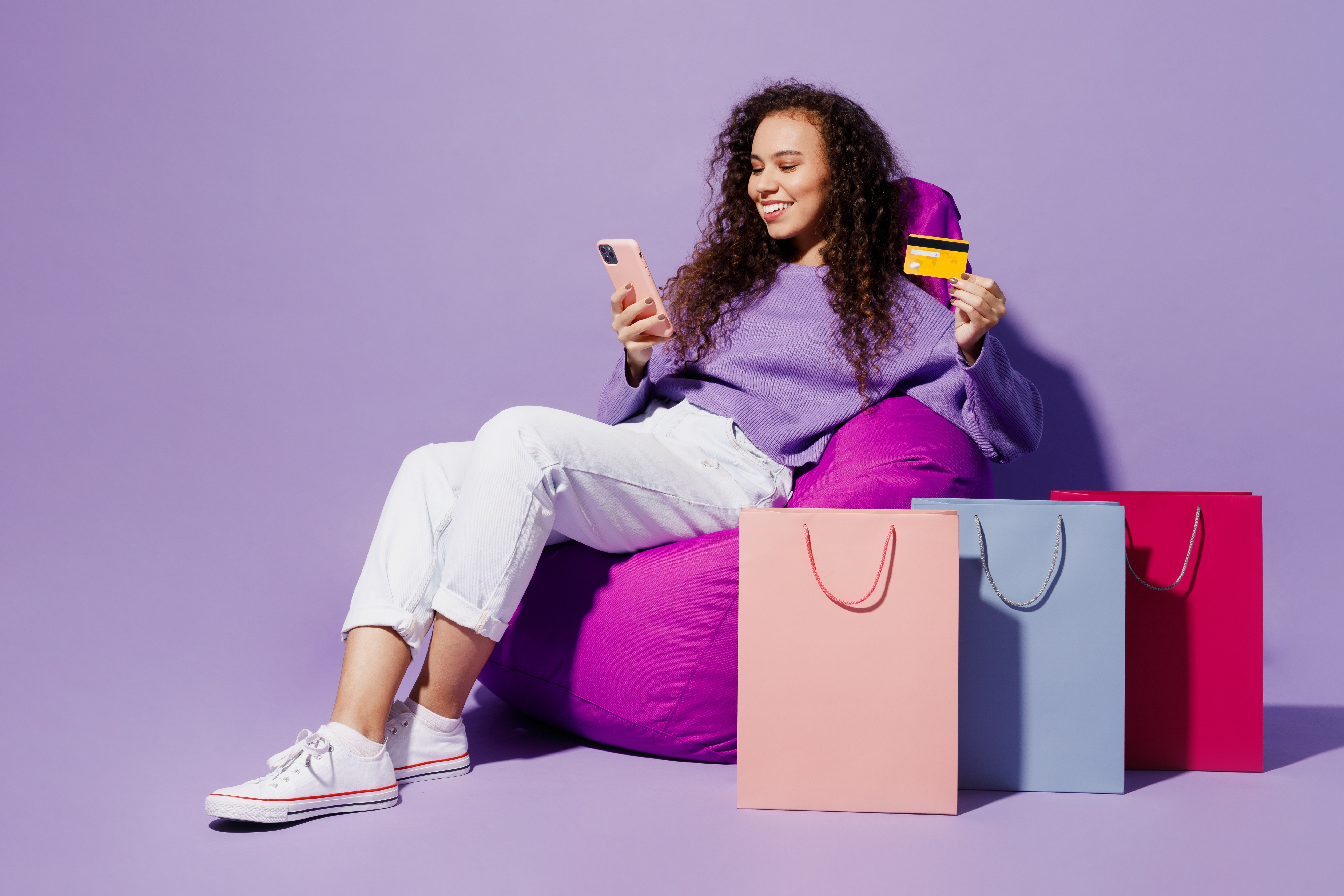 Une fille portant un pull s'assoit sur une chaise près des sacs d'emballage après le shopping, utilise une carte de crédit de téléphone portable isolée sur fond violet clair pastel uni.