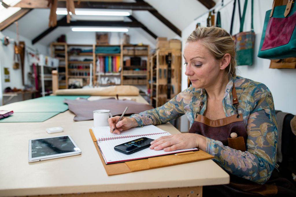 Une tanneuse assise dans son atelier de tannerie, à un établi, met à jour ses livres de comptes avec les bénéfices des ventes qu'elle a réalisées.